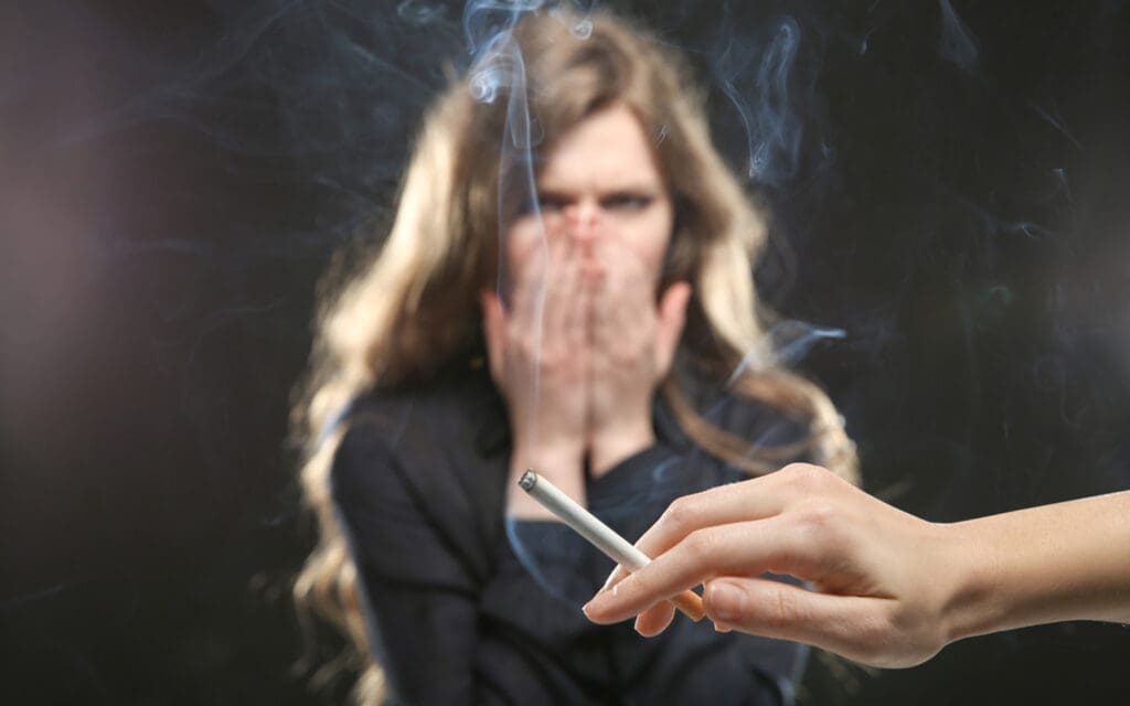 close shot of a hand holding a cigarette and a girl have both hands on her nose, seems like she is getting annoyed by cigarette smoke.