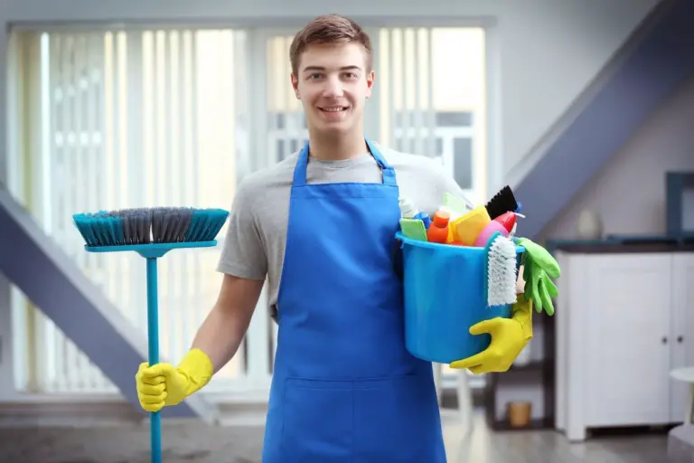 a professional cleaner in picture holding a brush in his right hand and a bucket with cleaning accessories in his left hand.