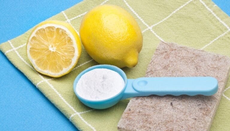 two lemons, one full and one half lemon, baking soda in a spoon, sponge under spoon and all these items are on a cleaning cloth for homemade cleaning remedies.